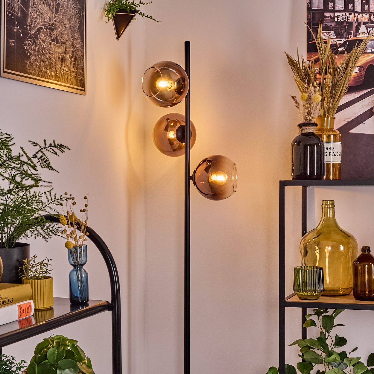 Eine moderne Stehlampe mit drei Glaskugeln. Regale mit Vasen, Pflanzen und Büchern. Kartenkunst an der Wand.