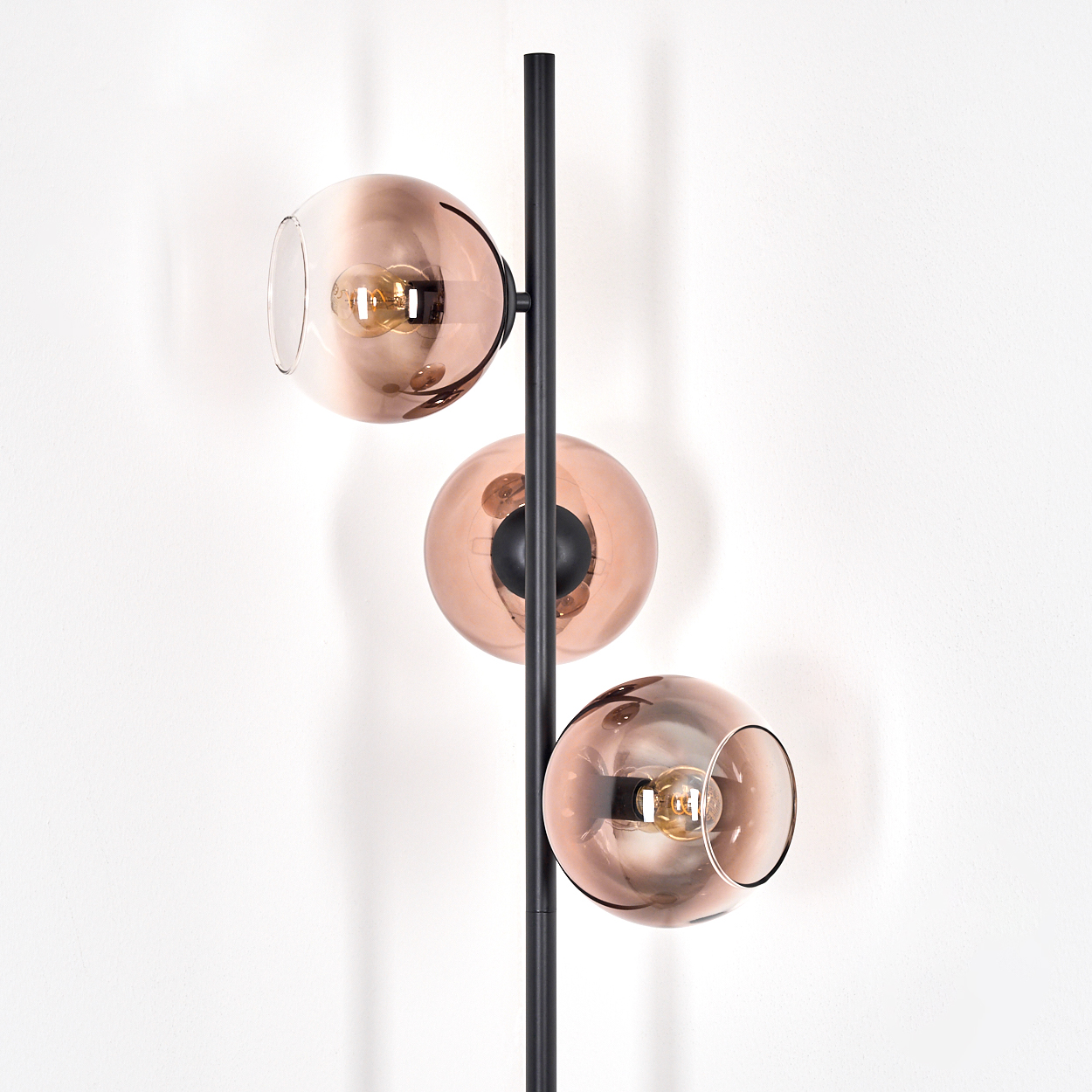 Eine moderne Stehlampe mit drei kugelförmigen, kupferfarbenen Glasschirmen, jede mit einer Glühbirne. Die Lampe steht auf einer schwarzen Stange vor einer weißen Wand.