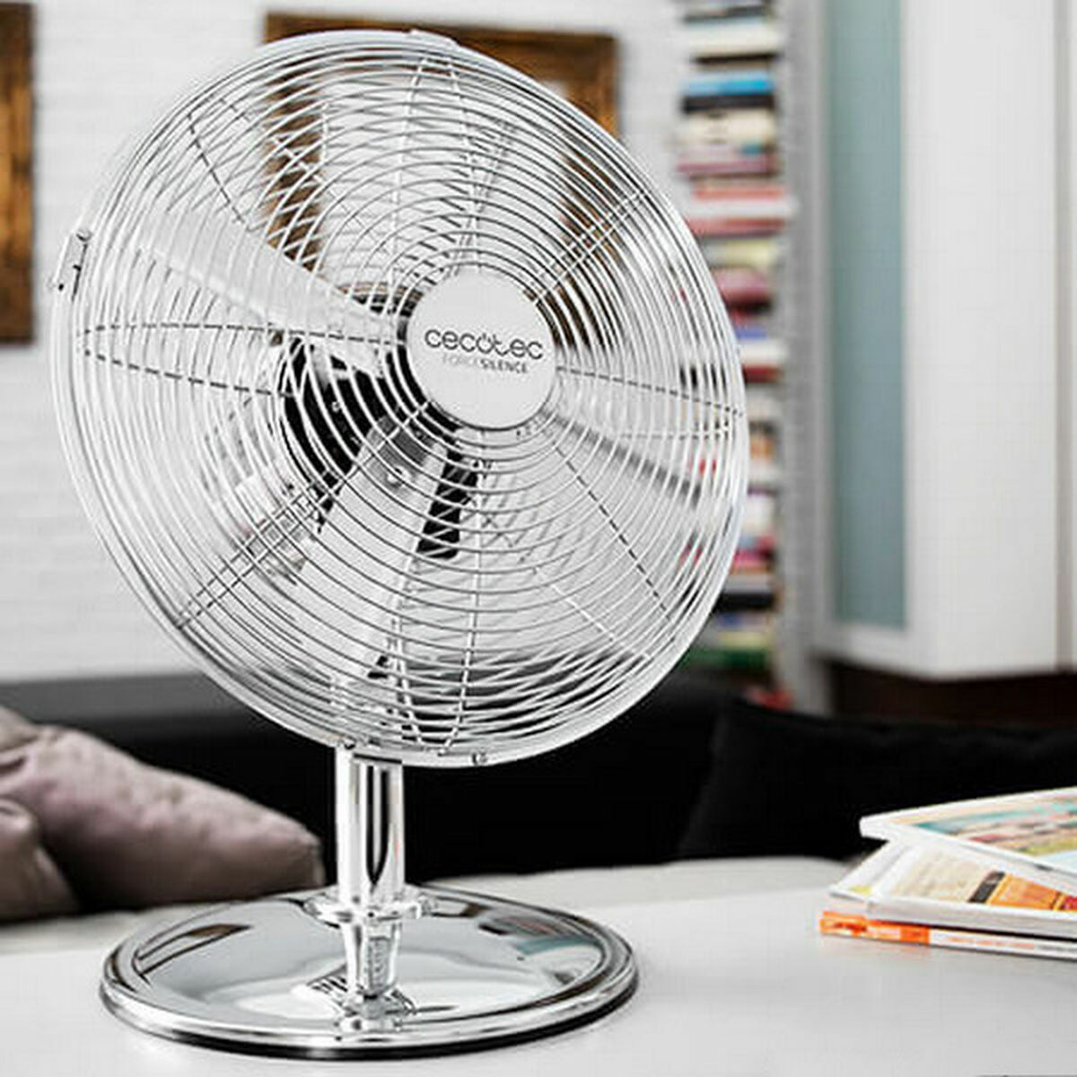 Un ventilador cromado está sobre una mesa. El ventilador está en primer plano, con un fondo borroso de una habitación, sofá y libros.
