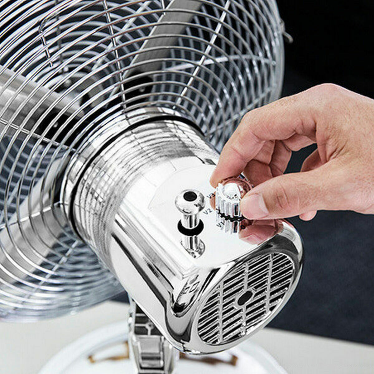 Una mano ajustando el dial de velocidad de un ventilador cromado. El ventilador es plateado con una jaula y una base pulida.