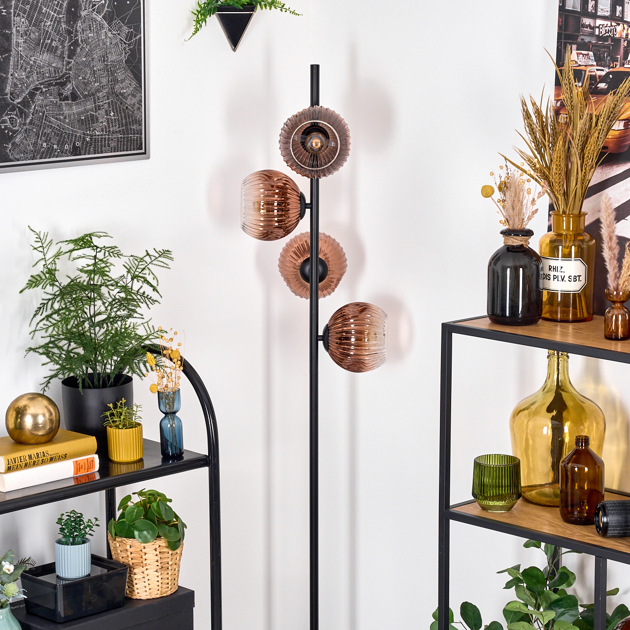 Ein Raum mit einem schwarzen Metallregal, einer Stehlampe, Pflanzen und dekorativen Glasflaschen. An der Wand hängt eine Karte.