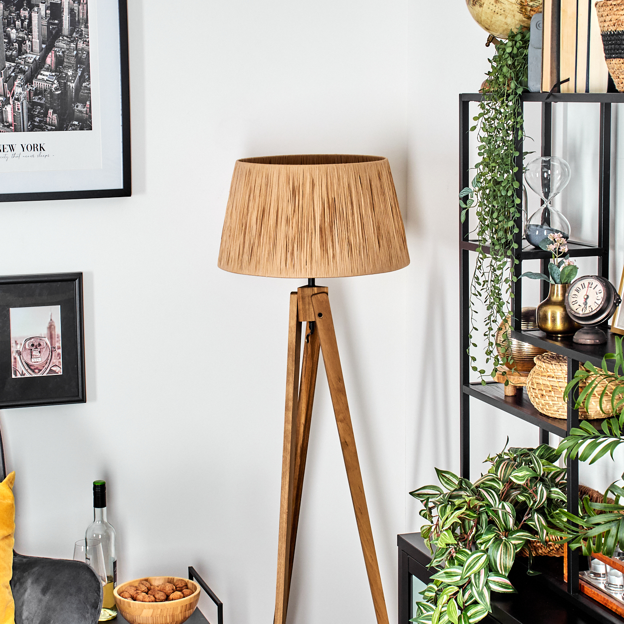 Ein Raum mit einer Holzlampe, schwarzen Regalen mit Pflanzen und Dekoration sowie Bildern an der weißen Wand. Eine Schale mit Nüssen.