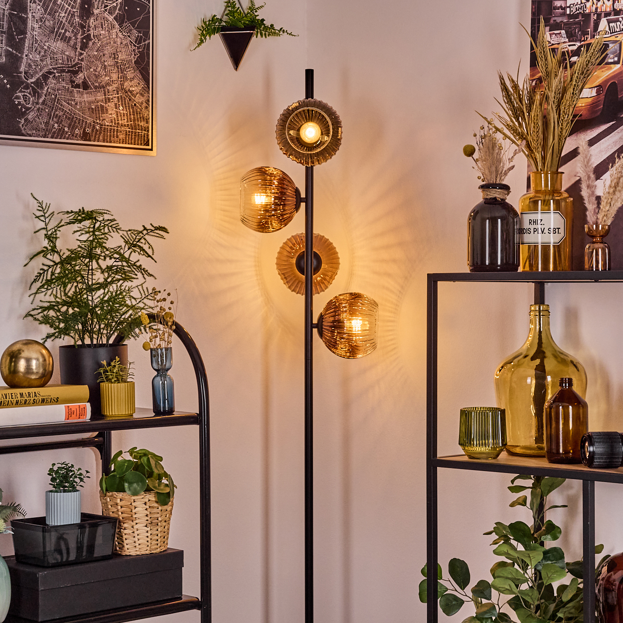 Ein Raum mit einem schwarzen Metallregal und einer Stehlampe. Auf dem Regal stehen Pflanzen, Vasen und Bücher. An der Wand hängt eine Karte.