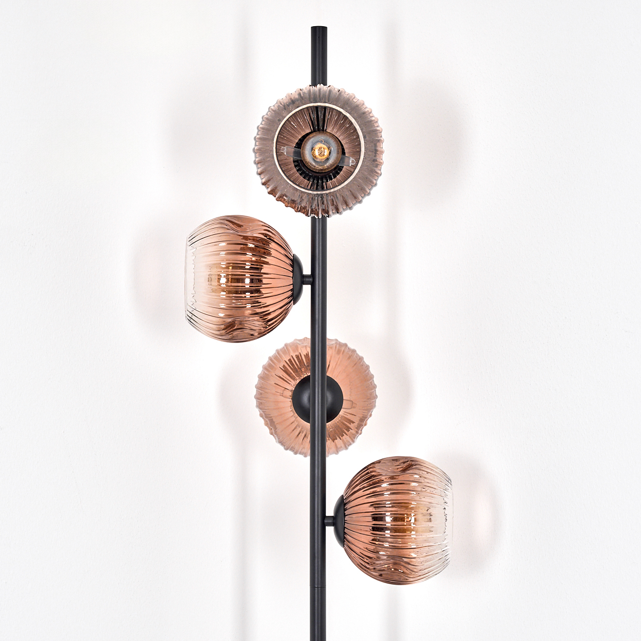 Eine moderne Lampe mit schwarzem Mast und drei dekorativen Glasschirmen in Braun- und Kupfertönen vor weißer Wand.