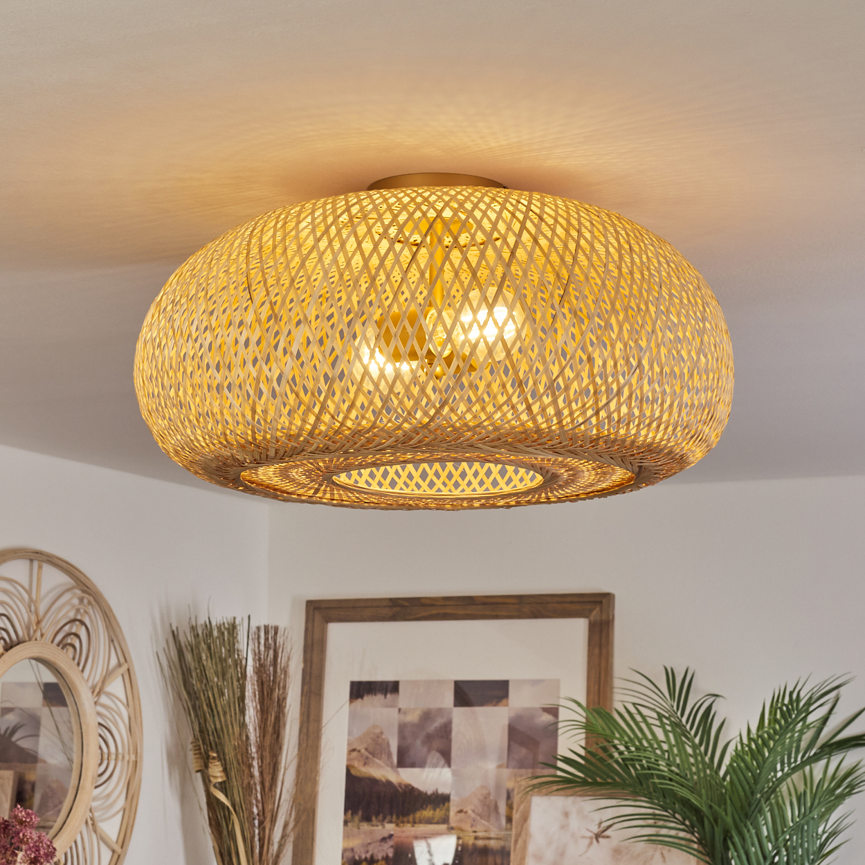Eine Deckenlampe aus gewebtem Bambus hängt in einem Raum. Darunter sind ein gerahmtes Bild und eine Pflanze zu sehen.