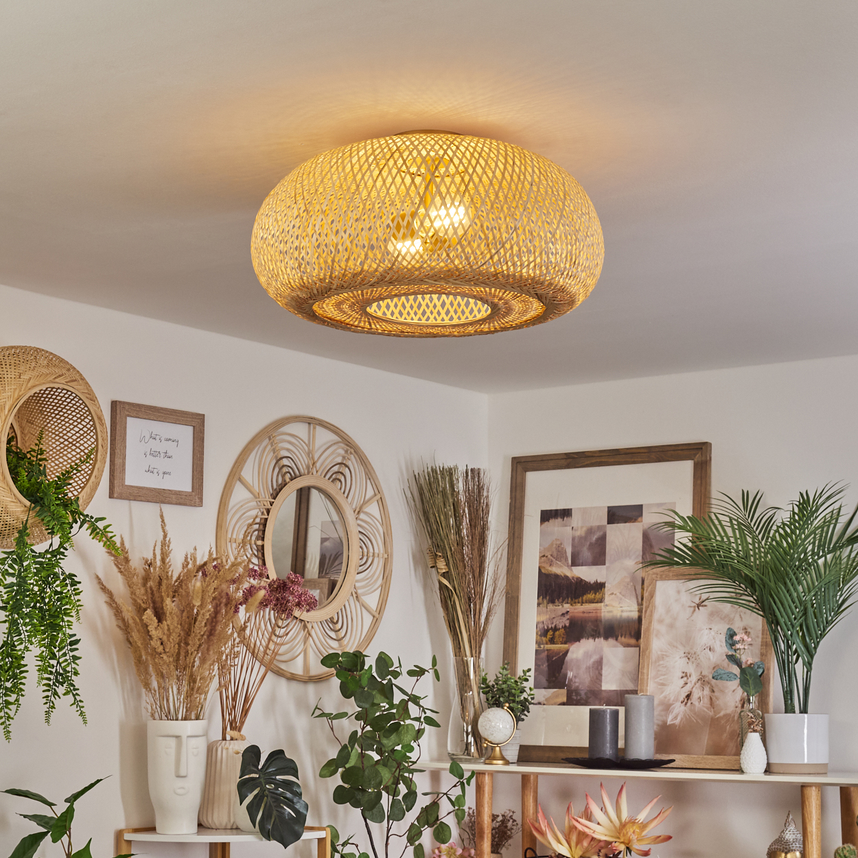 Eine gewebte Deckenlampe hängt über der Dekoration. Darunter schmücken Pflanzen, Spiegel und Bilder die weißen Wände und ein Regal.