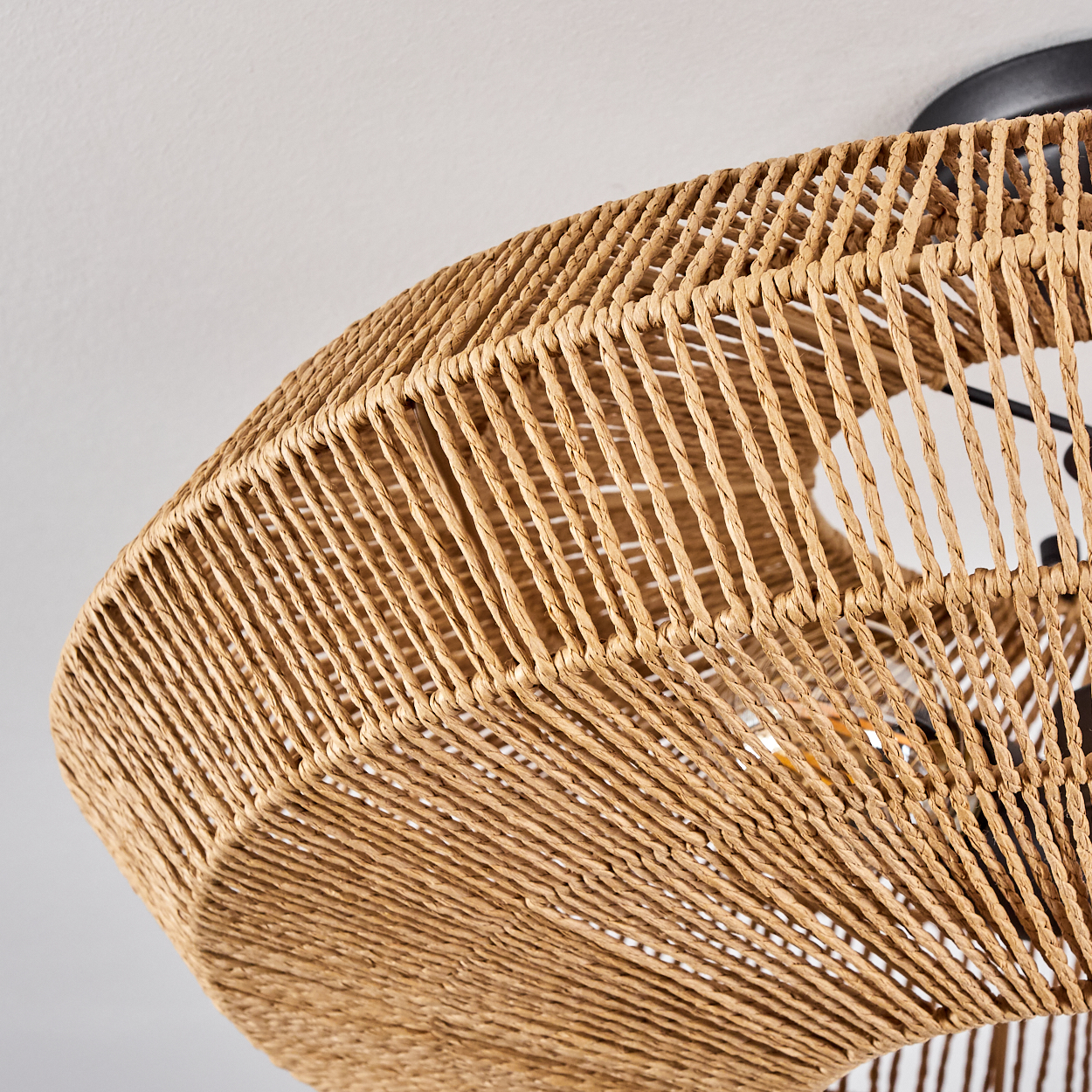 Nahaufnahme eines gewebten Korblampenschirms, hellbraun. Der Hintergrund ist eine weiße Decke. Die Lampe hängt.