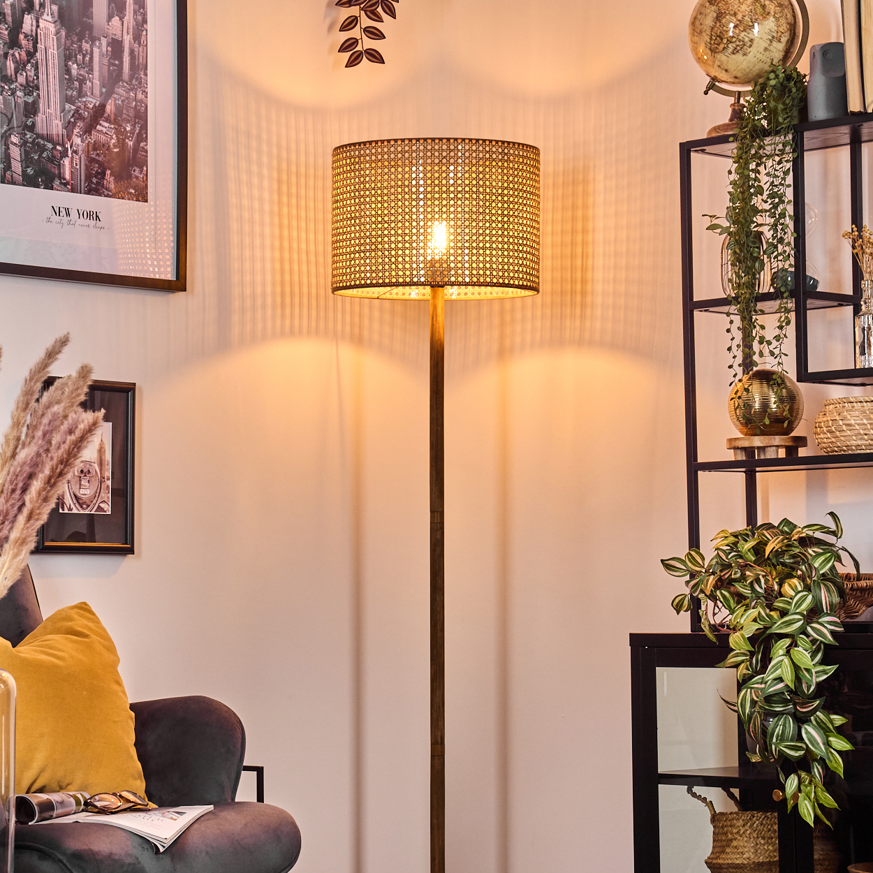 Eine beleuchtete Stehlampe mit gewebtem Schirm steht in einem Raum. Ein gerahmter New-York-Druck, ein Stuhl und ein Regal mit Dekoration sind ebenfalls sichtbar.