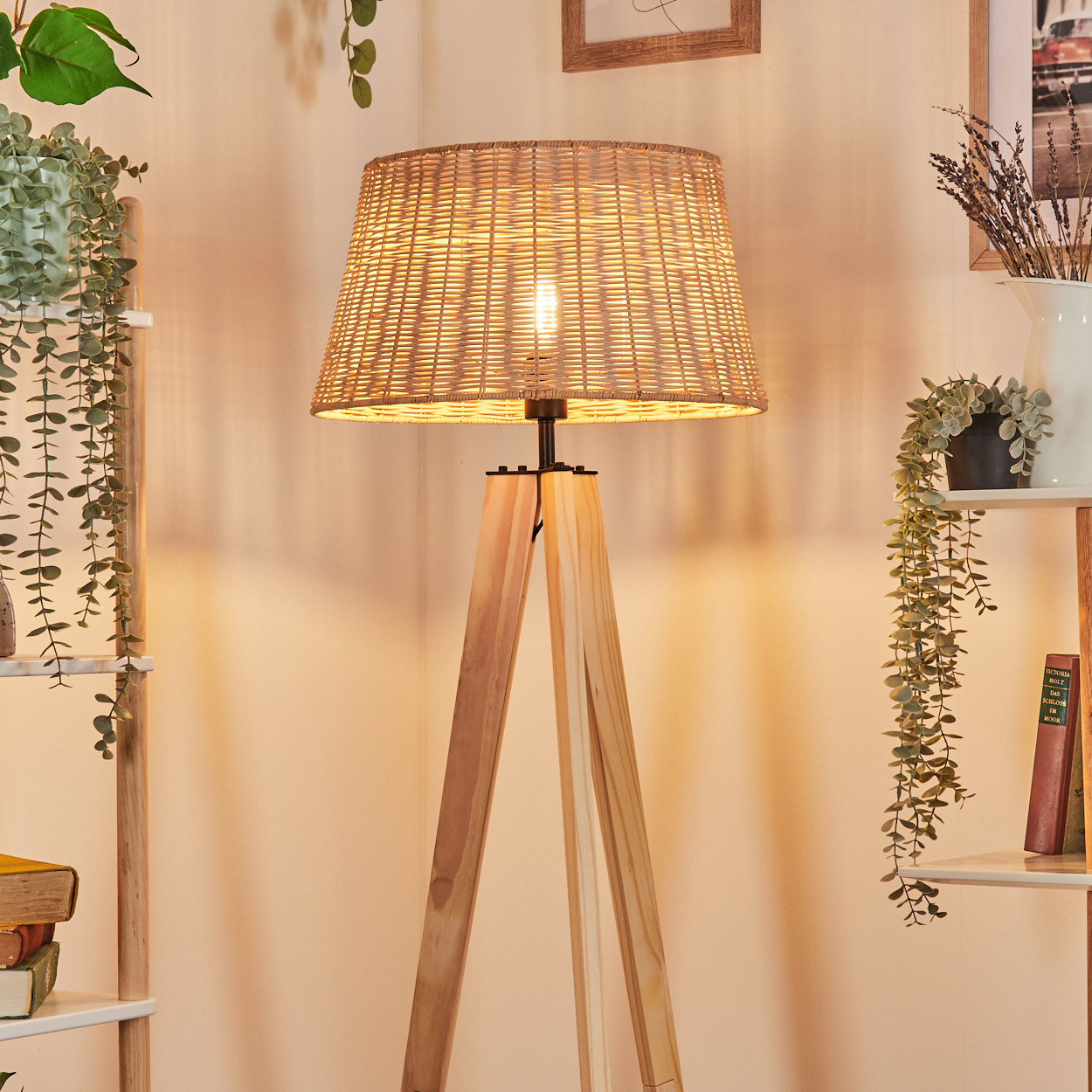 Eine Holzstativlampe mit gewebtem Lampenschirm ist beleuchtet. Pflanzen und Regale mit Büchern befinden sich auf beiden Seiten. Ein heller, warm getönter Raum.