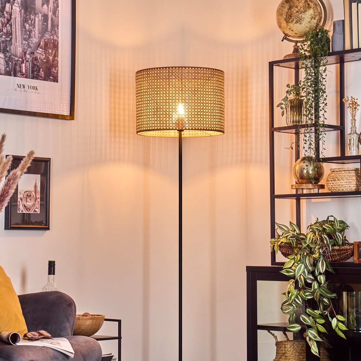 Eine beleuchtete Rattanlampe steht in einem Raum mit einem New York-Poster, Pflanzen und einem Regal. Ein gemütlicher Sessel ist im Vordergrund.