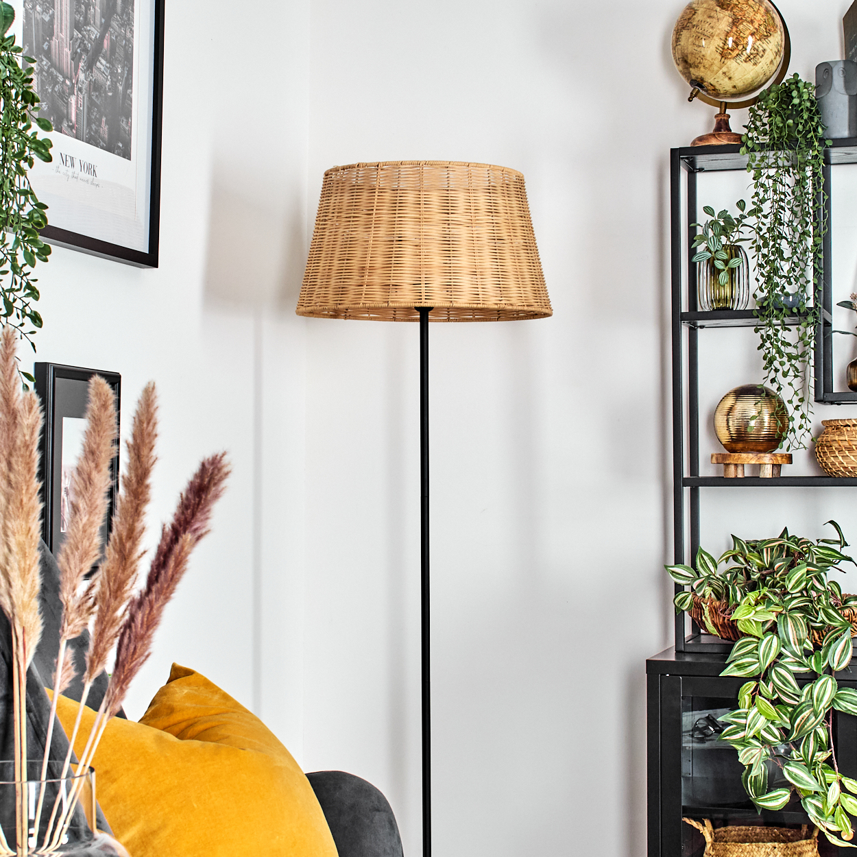 Eine Zimmerecke mit einer gewebten Lampe, schwarzen Regalen mit Pflanzen und Dekor. Ein gelbes Kissen und gerahmte Kunst sind sichtbar.
