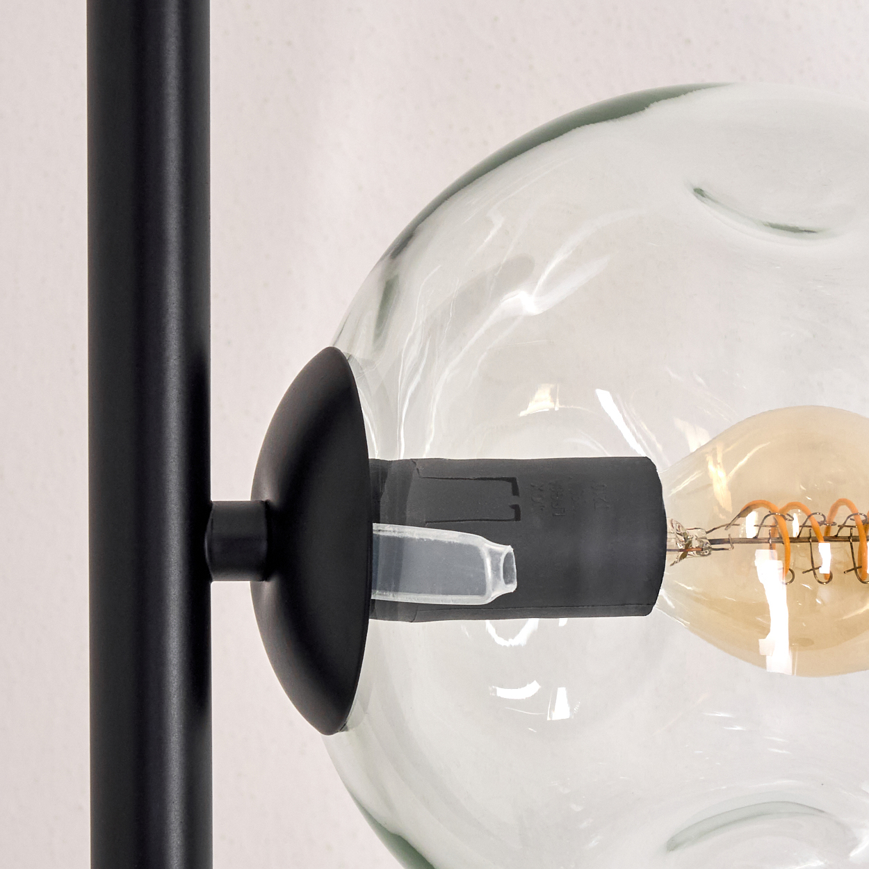 Nahaufnahme einer schwarzen Lampe mit Glaskugel und sichtbarer Glühbirne. Der Hintergrund ist eine schlichte weiße Wand.