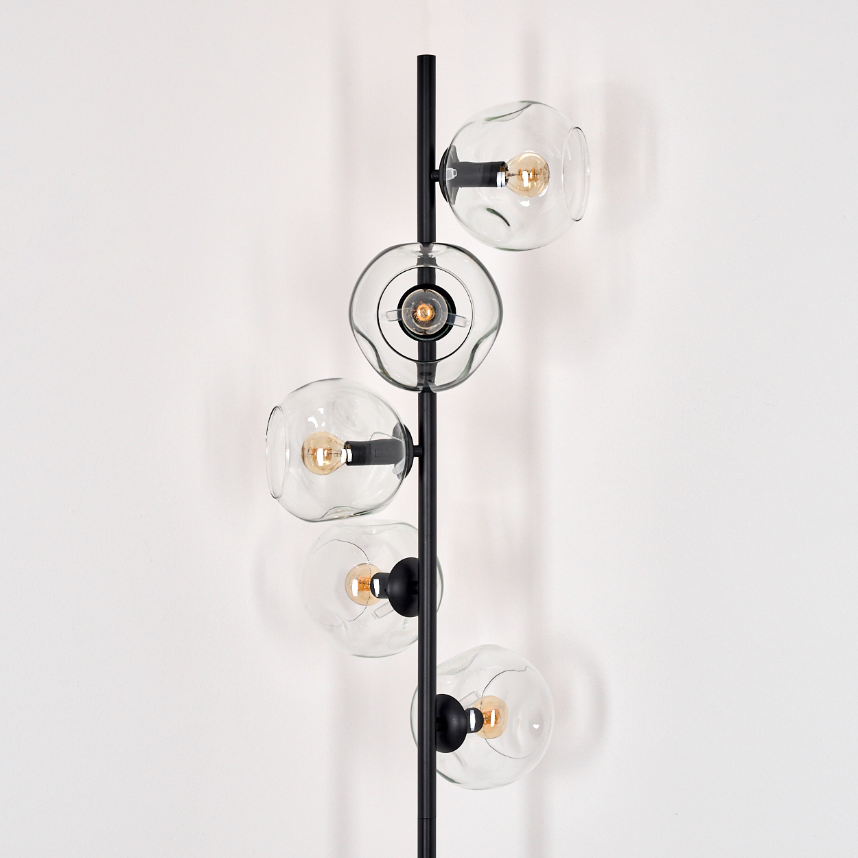 Eine moderne schwarze Stehlampe mit fünf klaren Glaskugeln, jede mit einer warmen Glühbirne. Sie steht vor einer weißen Wand.