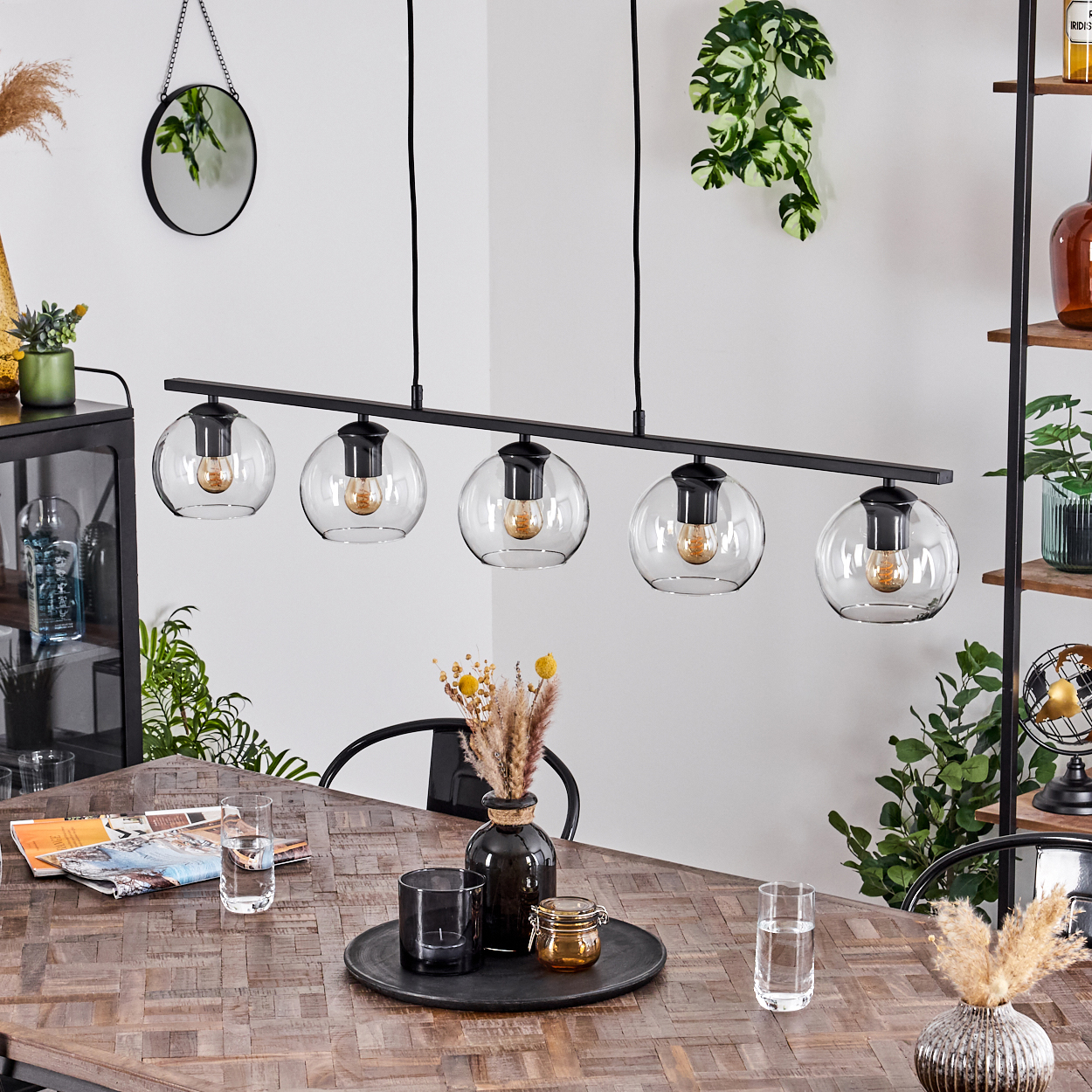 Ein Esszimmer mit Holztisch, schwarzem Kronleuchter und Dekoartikeln. Pflanzen und Spiegel an den Wänden.