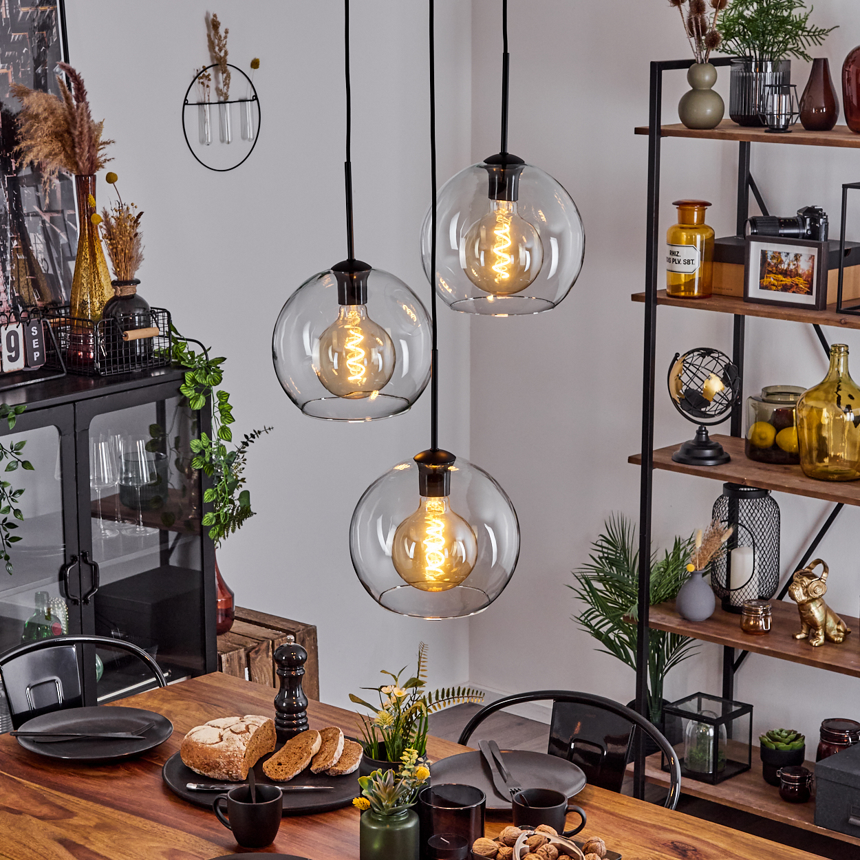 Esszimmer mit Holztisch, Hängelampen, schwarzen Tellern, Brot und dekorativen Regalen mit verschiedenen Gegenständen.