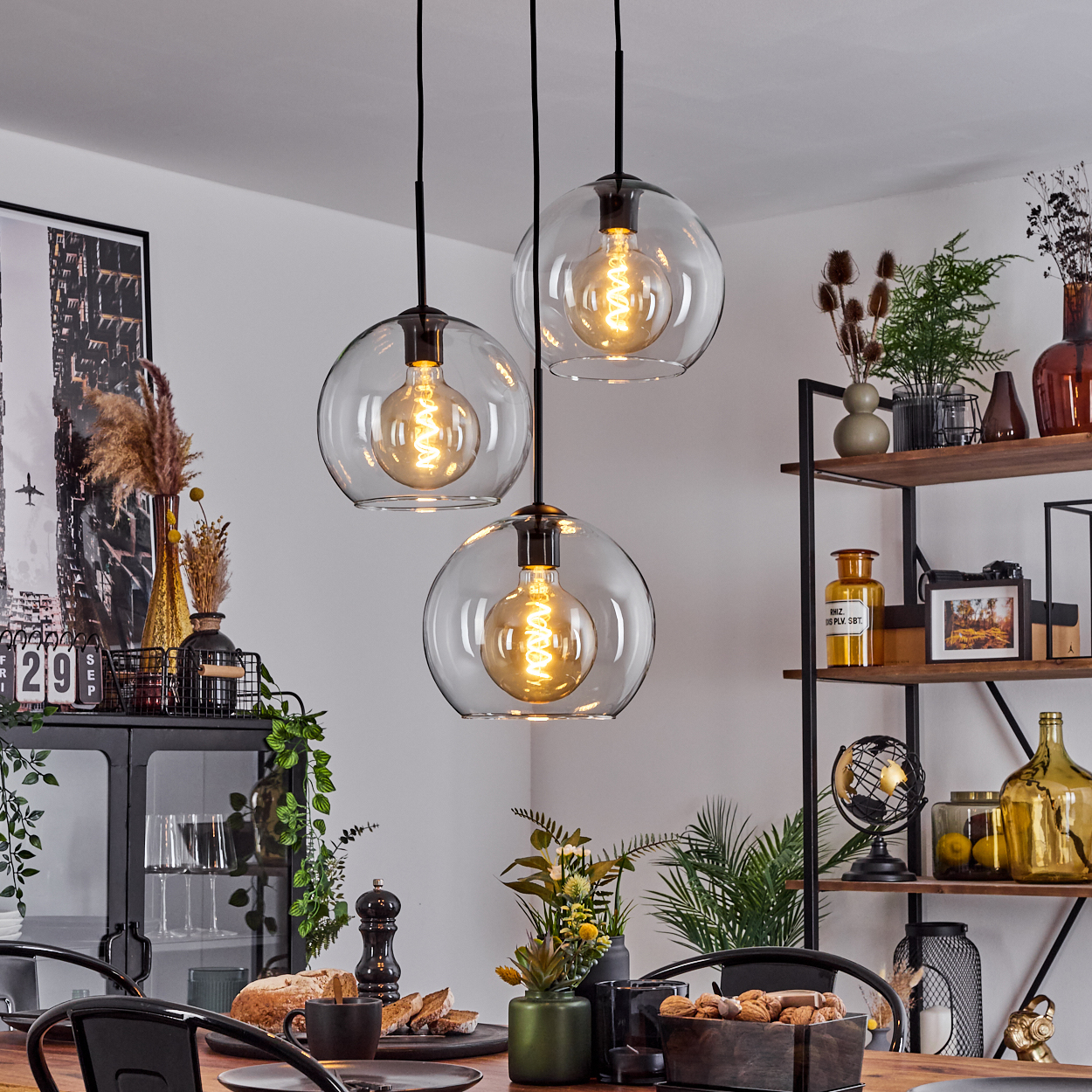 Drei Kugelhängeleuchten über einem Holztisch, der für eine Mahlzeit gedeckt ist. Regale mit Dekor befinden sich im Hintergrund.