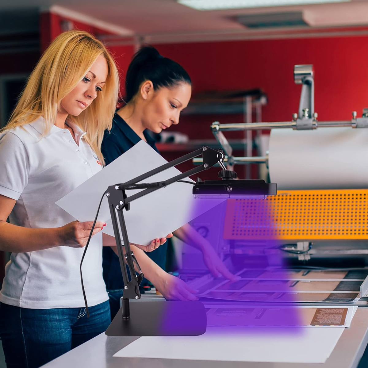 Zwei Frauen untersuchen in einer Druckerei Papiere unter einer schwarzen Lampe mit violettem Licht. Im Hintergrund steht eine Druckmaschine.