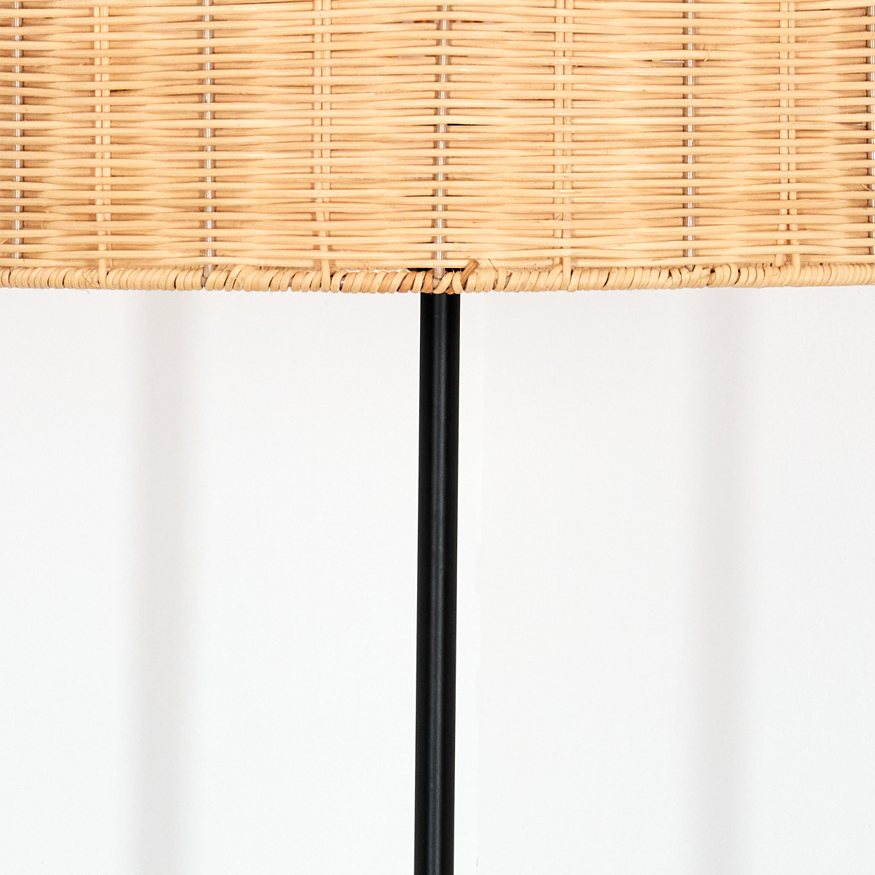 Nahaufnahme einer Lampe. Oben ein gewebter, hellbrauner Lampenschirm, in der Mitte ein schwarzer Pfosten, weißer Hintergrund.