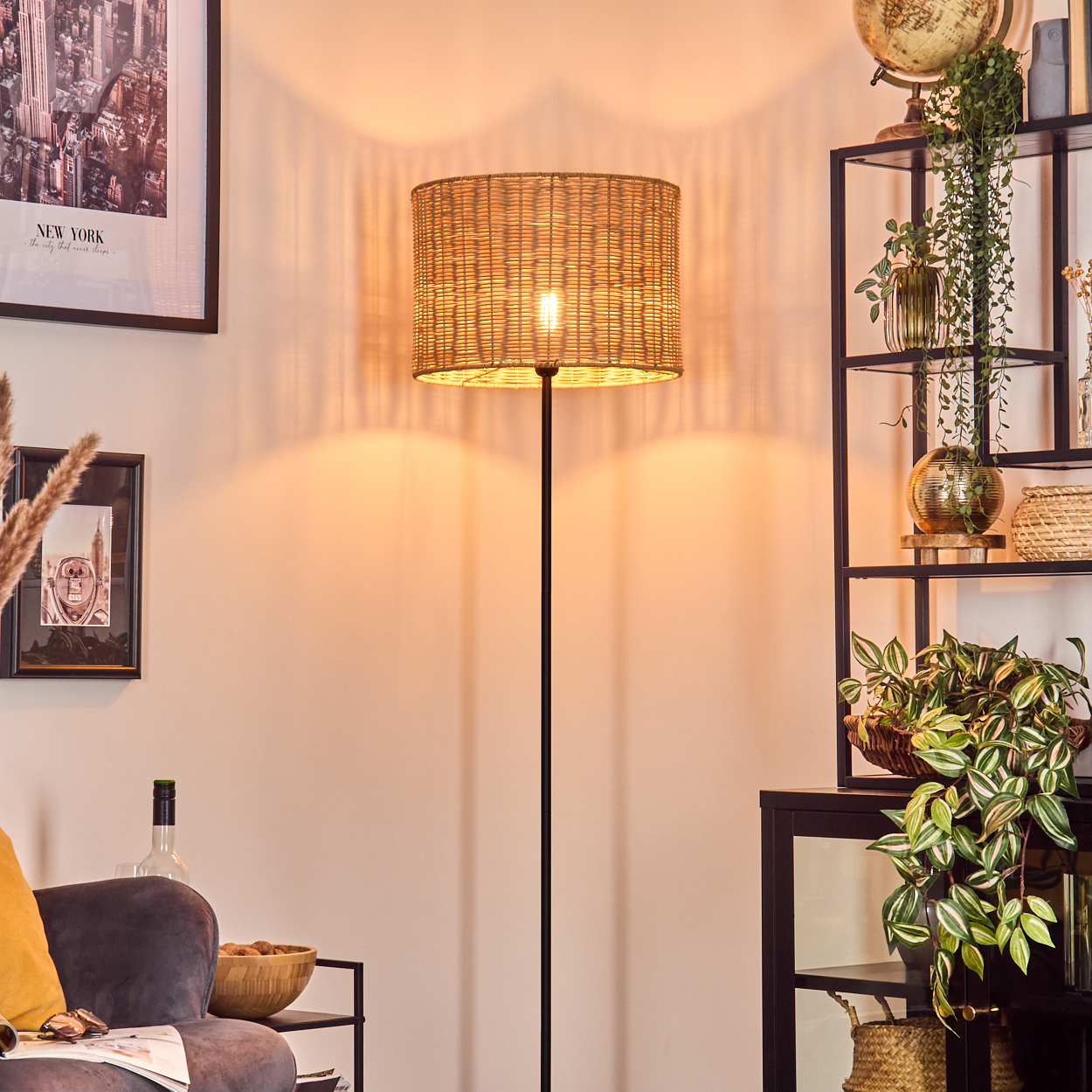 Eine beleuchtete Korblampe steht in einem Raum mit einem Regal voller Pflanzen, einem New York-Poster und einem Sofa mit gelbem Kissen.
