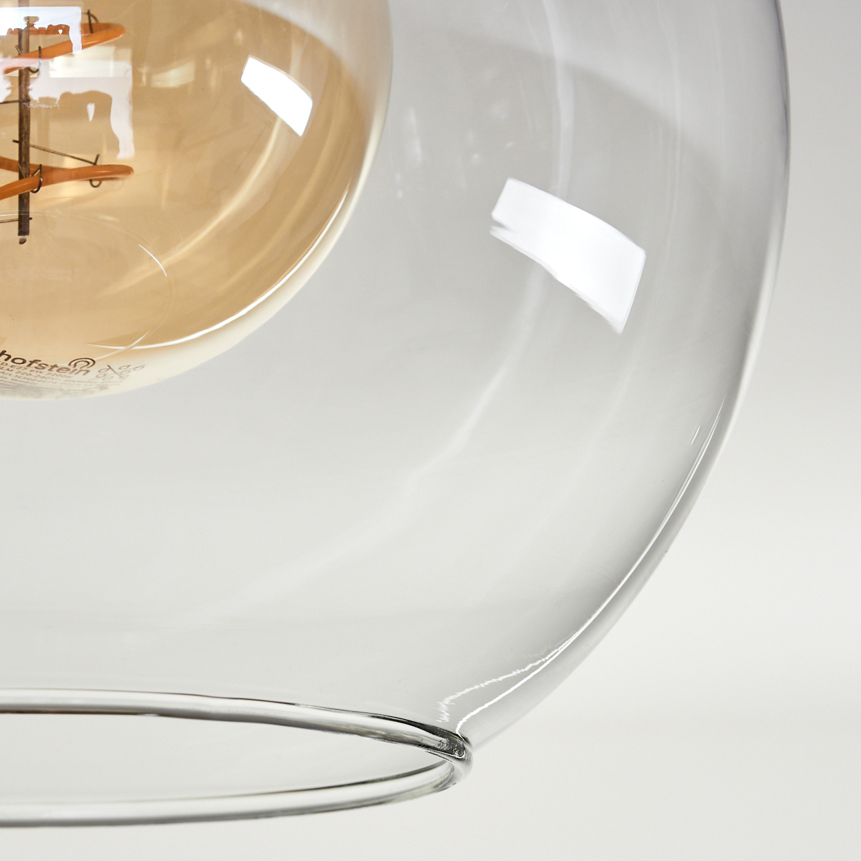 Ein klarer Glasschirm mit einer warm getönten Glühbirne im Inneren. Das Glas hat ein gebogenes Design und reflektiert Licht. Weißer Hintergrund.
