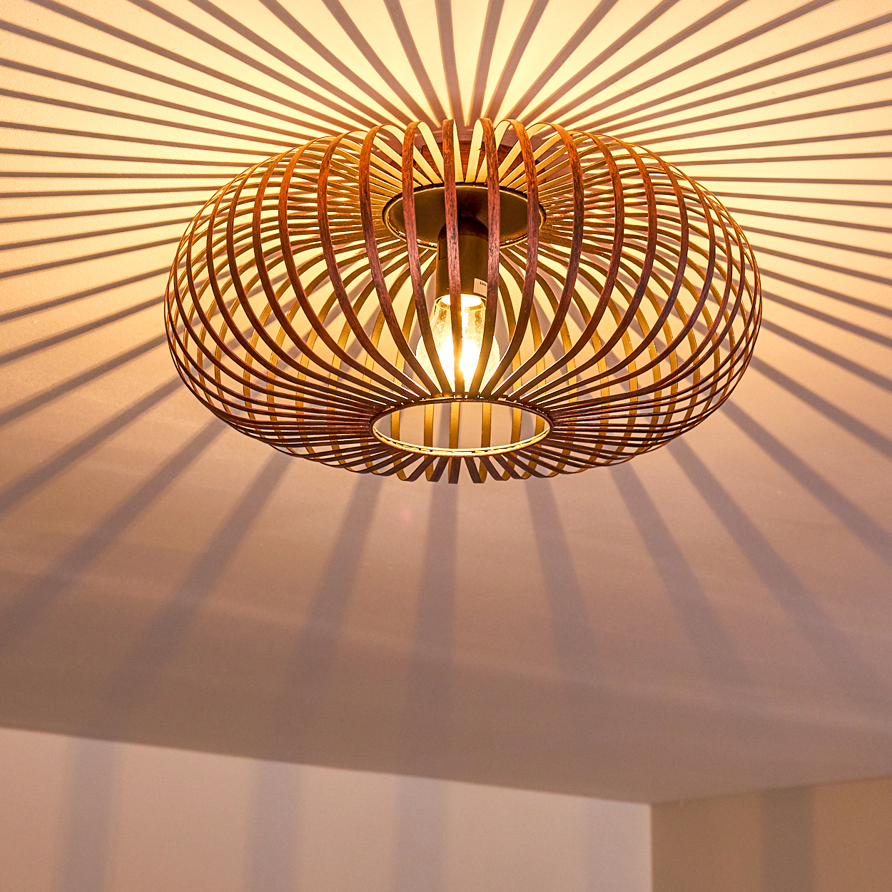 Eine dekorative Leuchte mit Holzgestell hängt von der Decke. Ihr Licht wirft Schatten. Die Glühbirne leuchtet.
