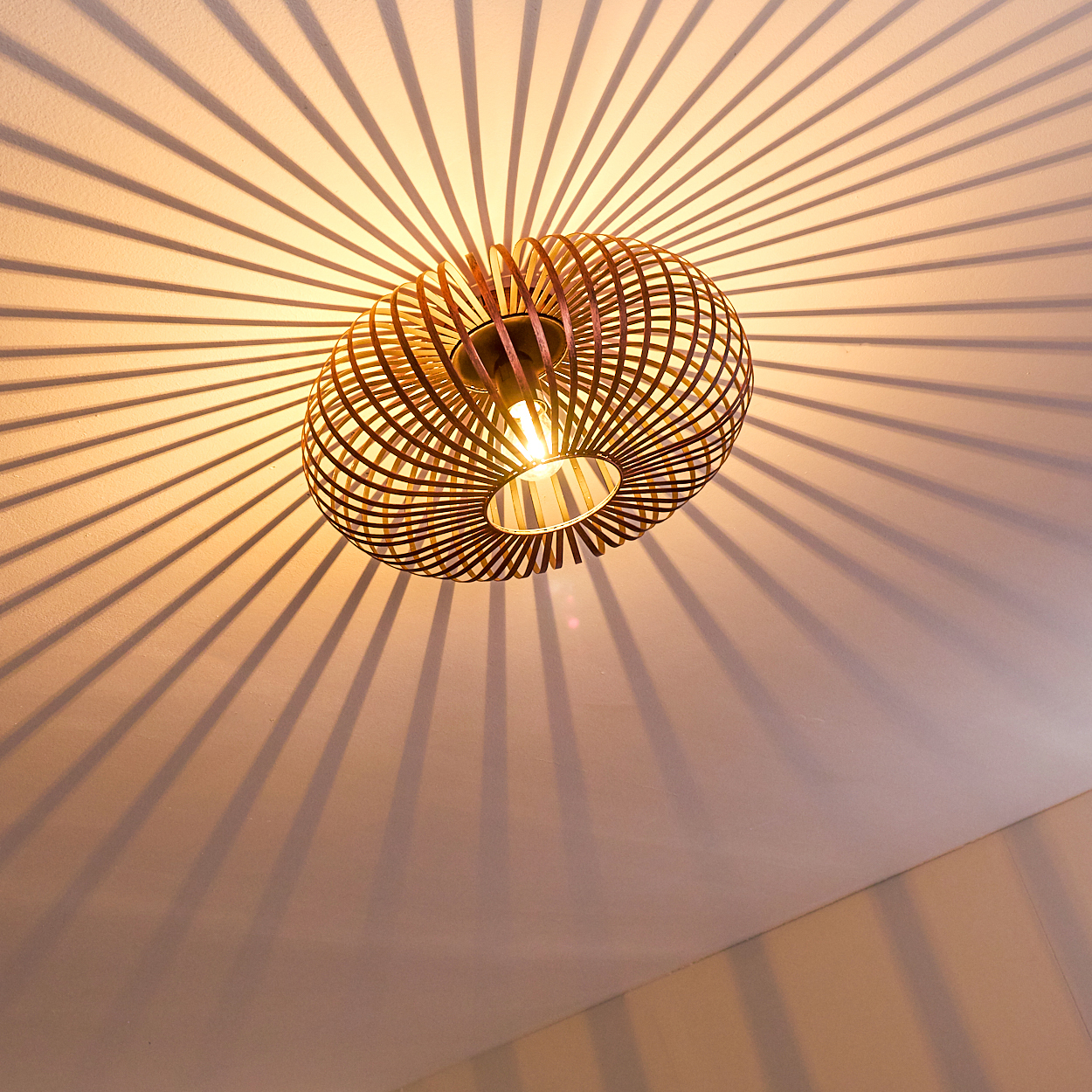 Eine dekorative Deckenlampe mit warmem Licht, die radiale Schatten auf die Decke wirft. Die Lampe besteht aus Holzstreifen.