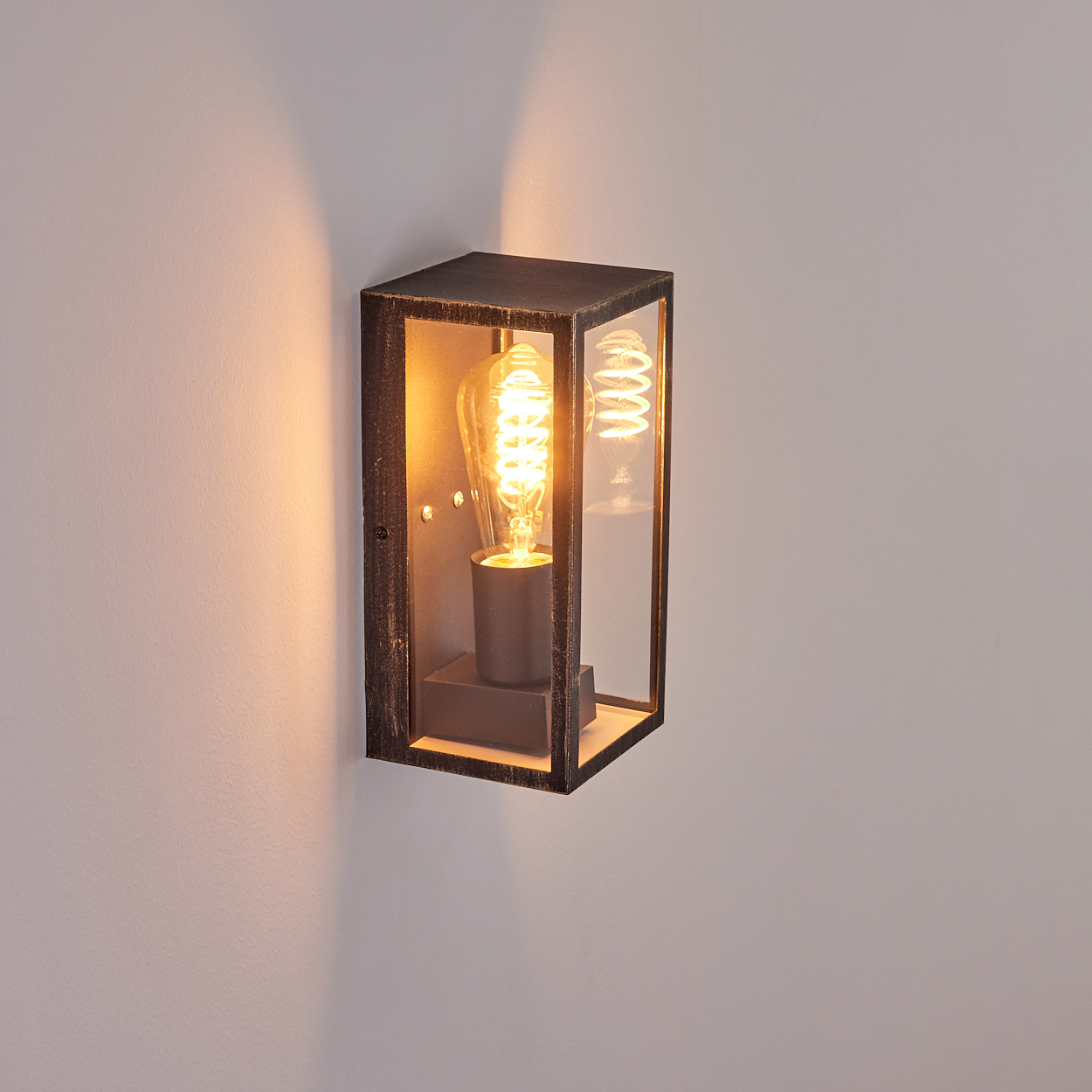 Eine rechteckige Außenwandleuchte. Sie hat ein Glasgehäuse und eine Glühbirne im Vintage-Stil. Die Leuchte ist an einer weißen Wand befestigt.