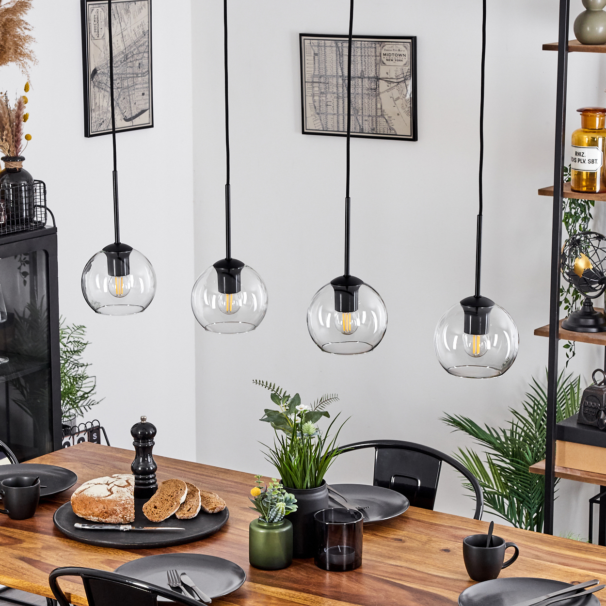 Esszimmer mit Holztisch, vier Pendelleuchten, Brot, Pflanzen und schwarzen Stühlen. Karten an der Wand und Regale mit Gegenständen.