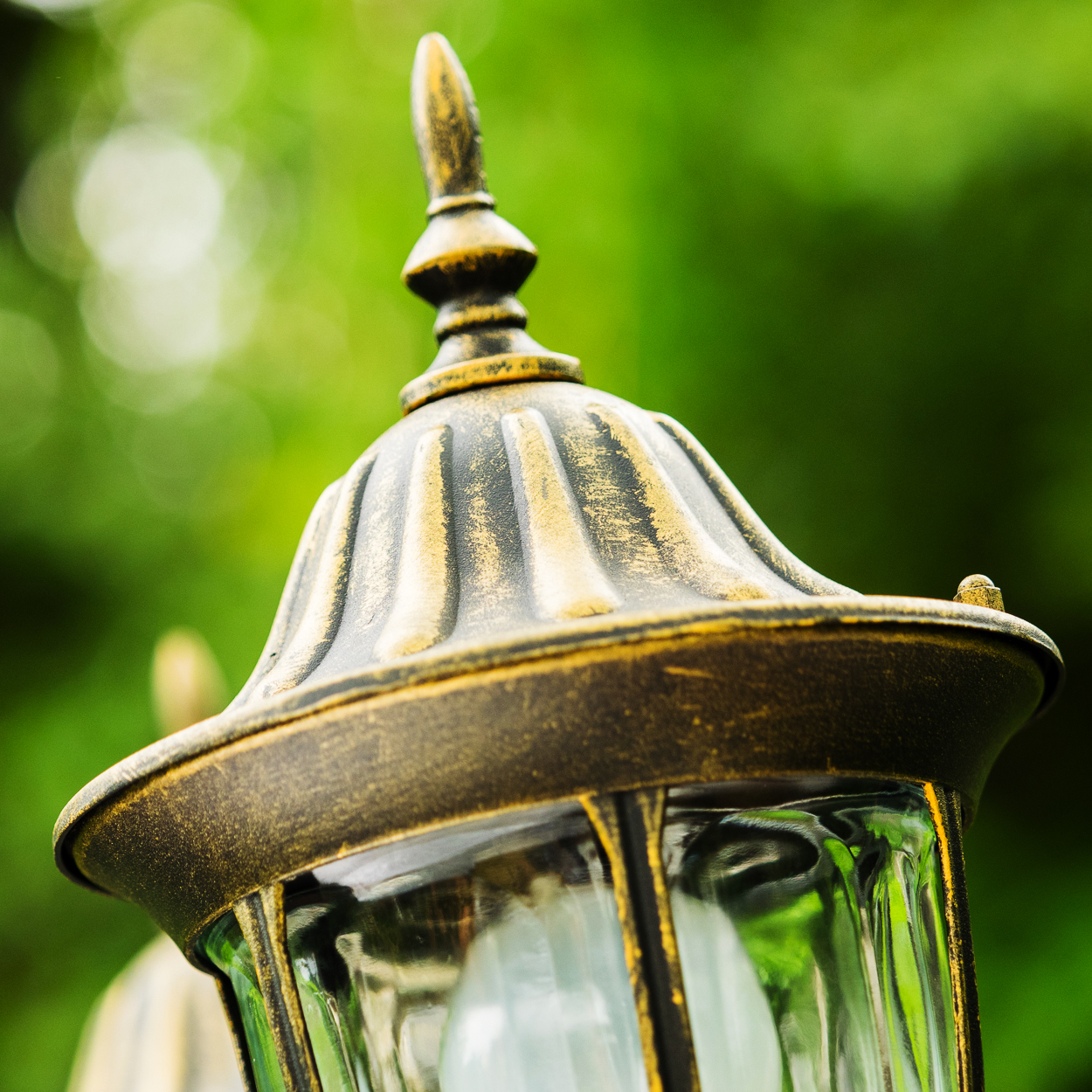 Nahaufnahme einer dekorativen Lampe mit Bronze-Finish. Grünes Laub im unscharfen Hintergrund. Die Lampe ist eine Gartenleuchte.