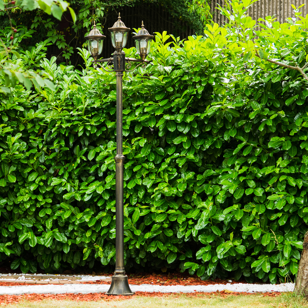 Eine dekorative Laterne mit drei Lichtern steht vor einer grünen Hecke. Der Boden ist mit Mulch und weißen Steinen bedeckt.