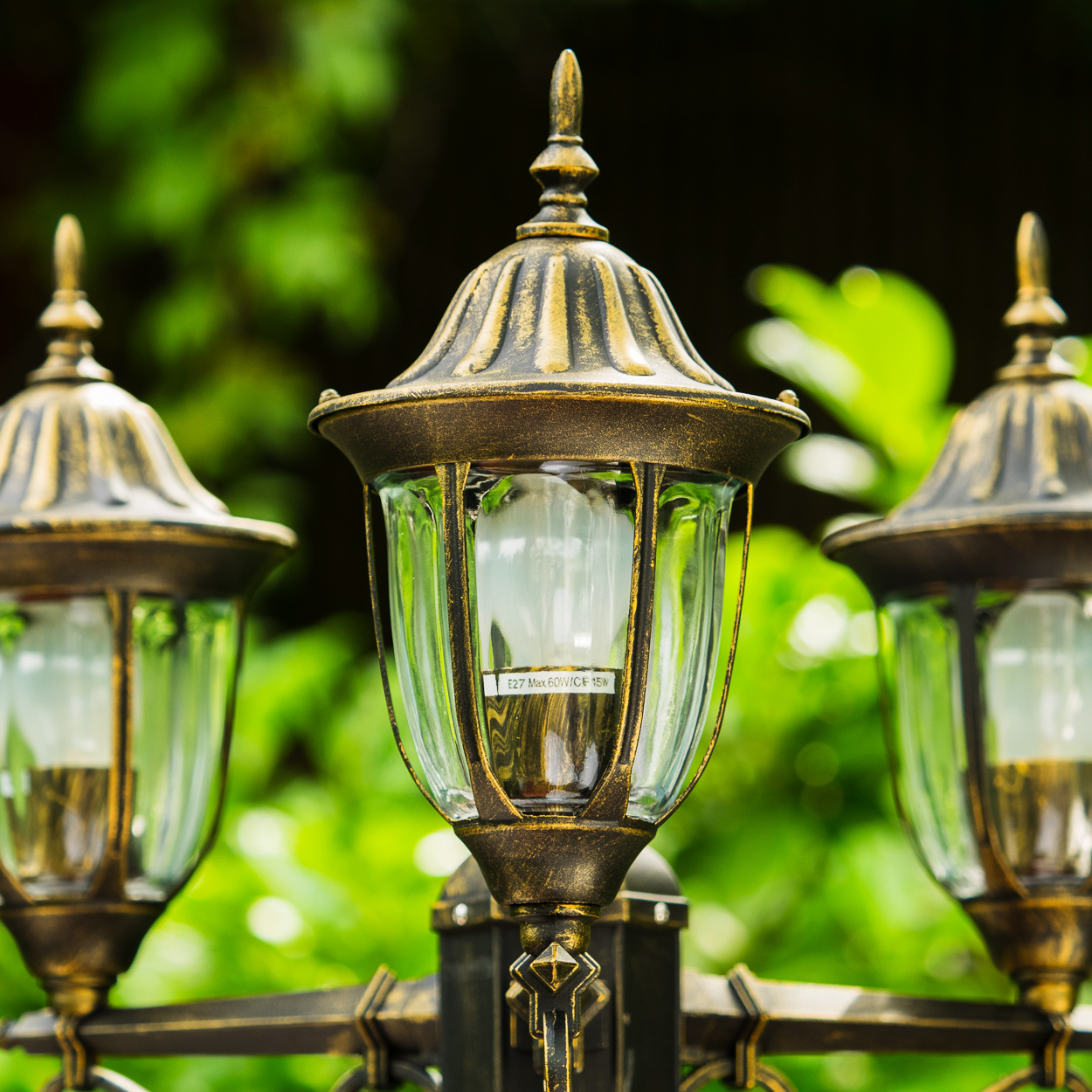 Drei verzierte, bronzefarbene Außenlampen mit Glasscheiben. Im Hintergrund ist grünes Laub zu sehen.