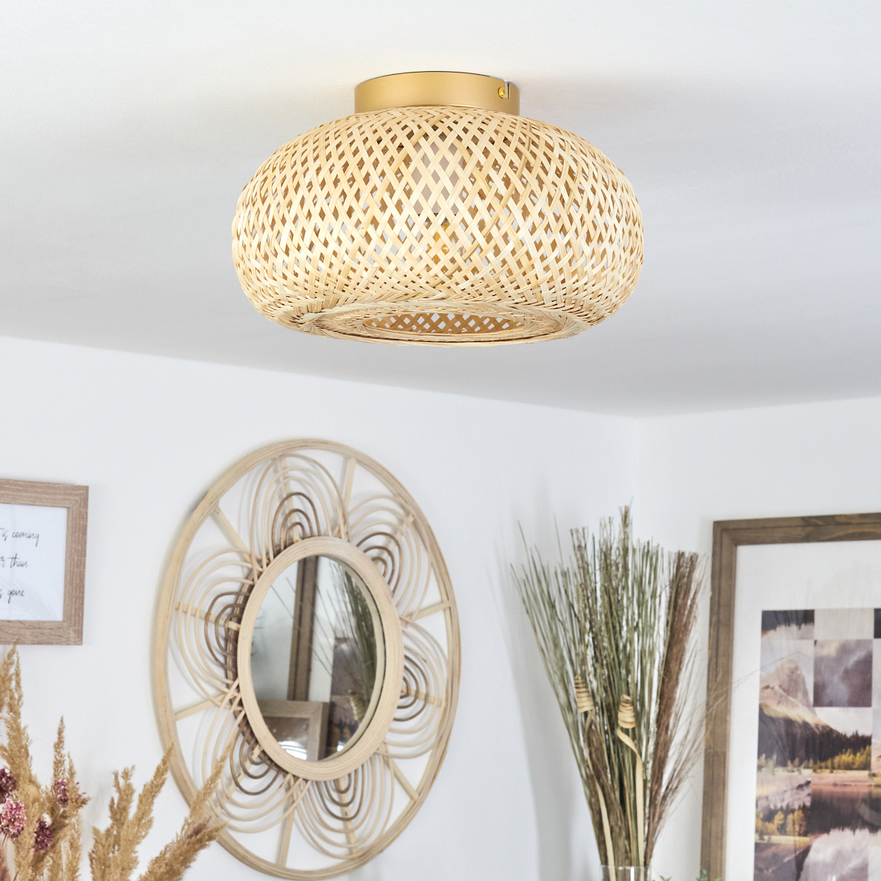 Eine Deckenlampe aus gewebtem Bambus, ein runder Spiegel und gerahmte Bilder schmücken eine weiße Wand. Trockene Pflanzen sind im Vordergrund.