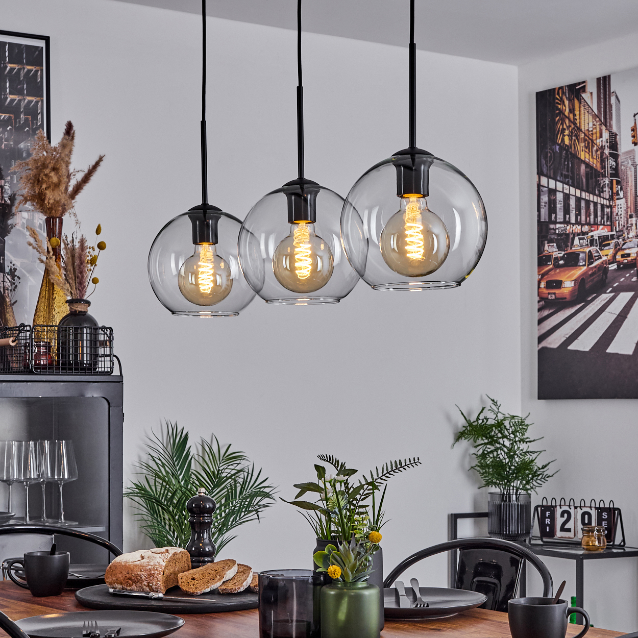 Esszimmer mit drei Hängelampen. Tisch mit Brot, Tellern und Blumen. Kunstwerk an der Wand mit einer Stadtszene.
