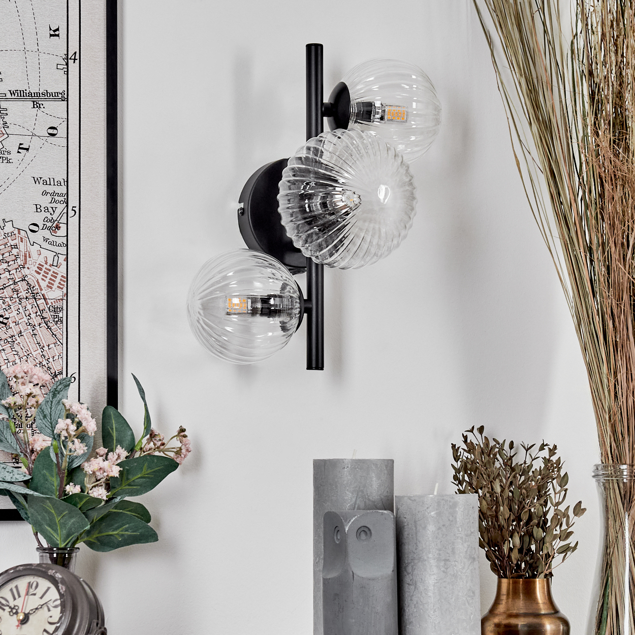 Eine moderne Wandlampe mit drei Glaskugeln. Hintergrund: Karte, Blumen, Kerzen und getrocknete Pflanzen in einer Vase.