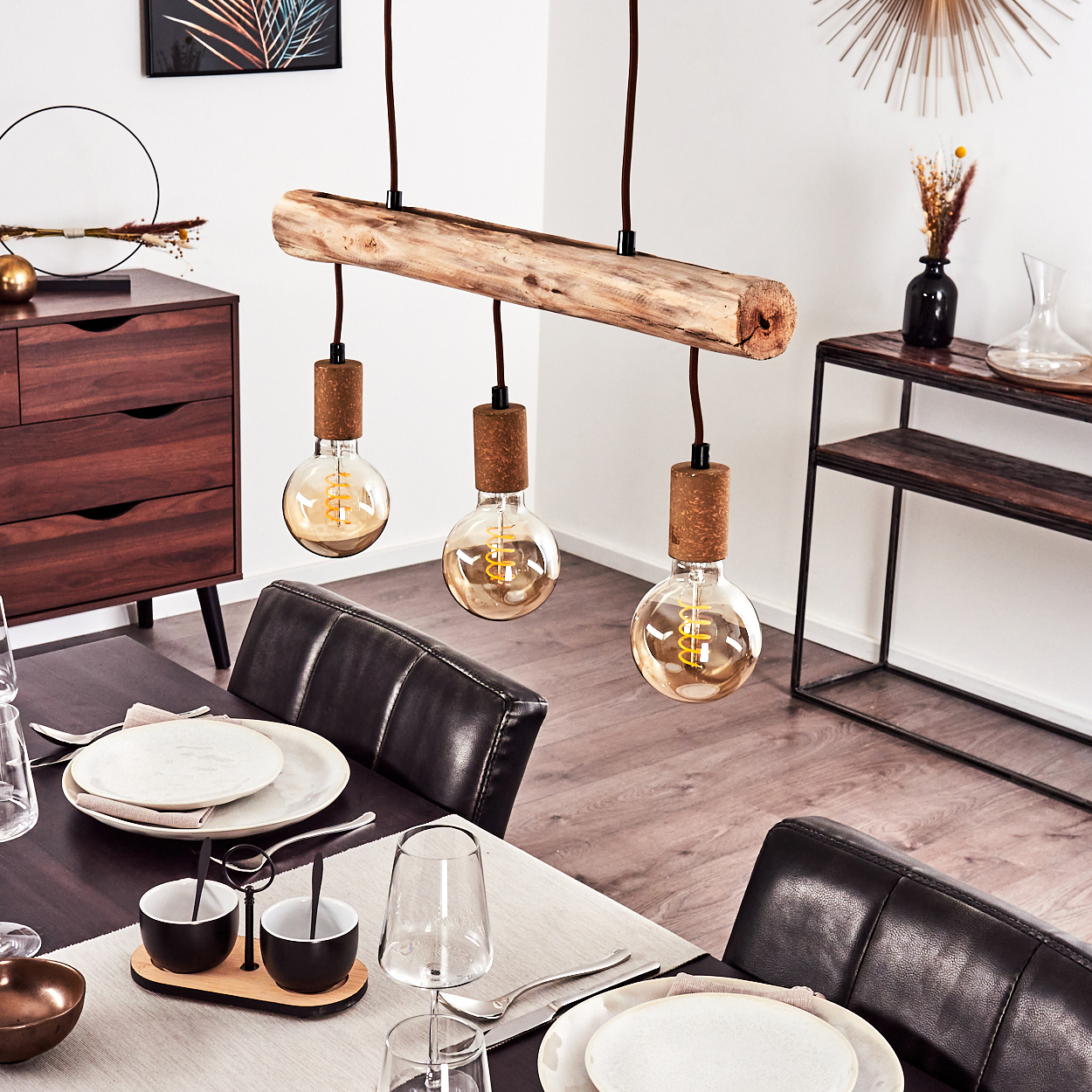 Ein modernes Esszimmer mit einer Holzbalken-Leuchte mit drei freiliegenden Glühbirnen, Tischdeko und einem dunklen Holzschrank.