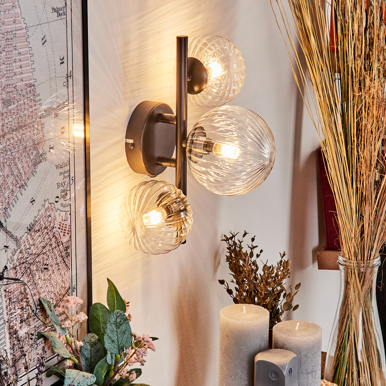 Eine Wandlampe mit drei Glaskugeln. Daneben sind eine Karte, Blumen, Kerzen und getrocknete Pflanzen angeordnet.