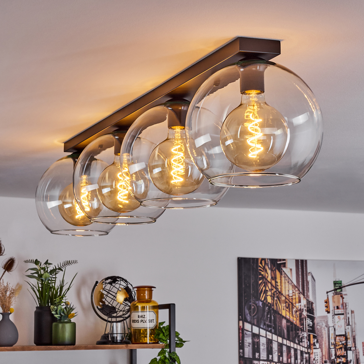 Moderne Deckenleuchte mit fünf großen Glaskugelschirmen, jede mit einer warm leuchtenden Glühbirne. Darunter ein Regal mit Dekoration.