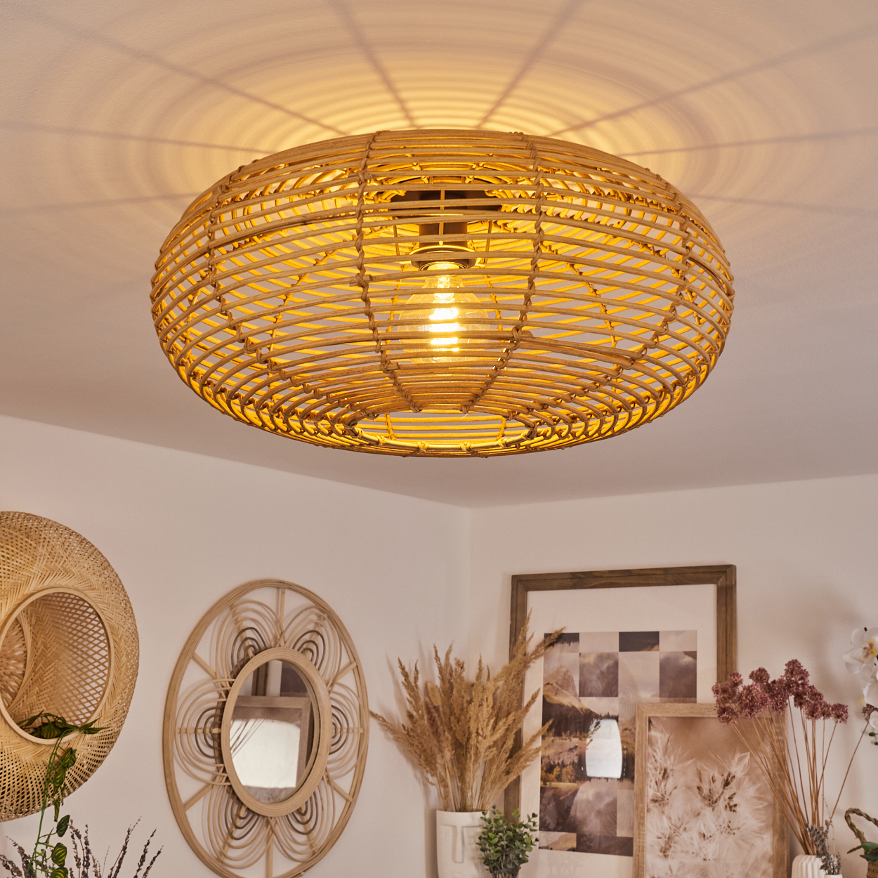 Eine gewebte Rattanlampe hängt von der Decke. Dekorative Spiegel und gerahmte Bilder befinden sich an der weißen Wand. Getrocknete Pflanzen sind in einer Vase.
