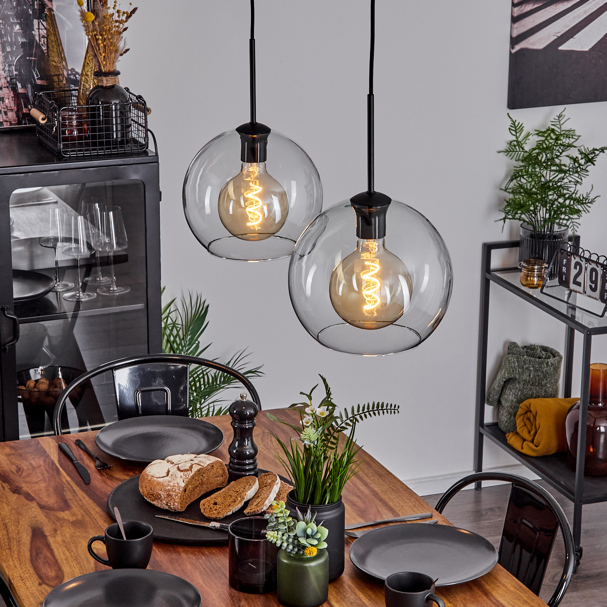 Esstisch mit Brot, Tellern und Blumen. Zwei Pendelleuchten hängen darüber. Ein Schrank und ein Metallregal sind im Hintergrund.