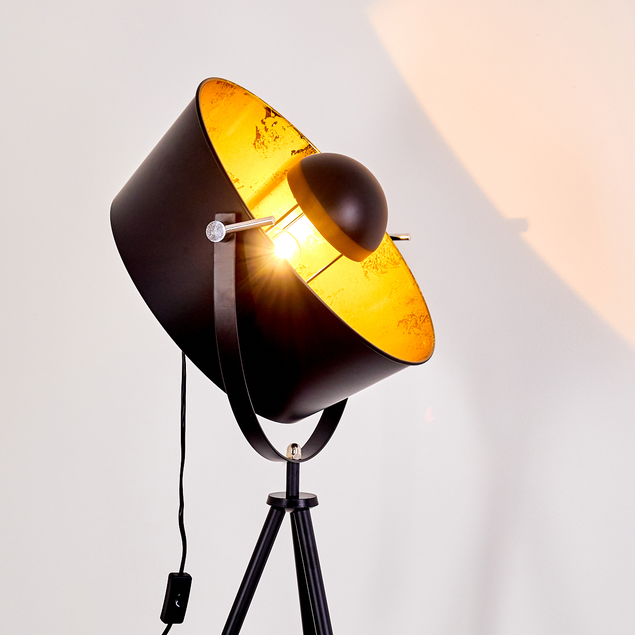 Eine schwarz-goldene Stehlampe mit Dreibein. Die Lampe ist abgewinkelt und wirft Licht auf eine weiße Wand. Das Licht ist an.