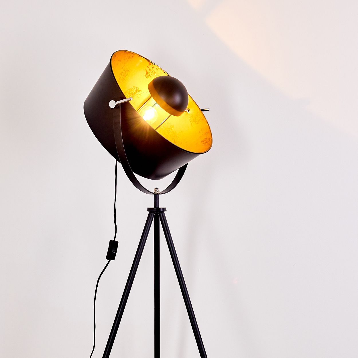 Eine schwarz-goldene Stehlampe mit Scheinwerferdesign. Die Lampe steht auf einem Stativ und der Hintergrund ist eine weiße Wand.