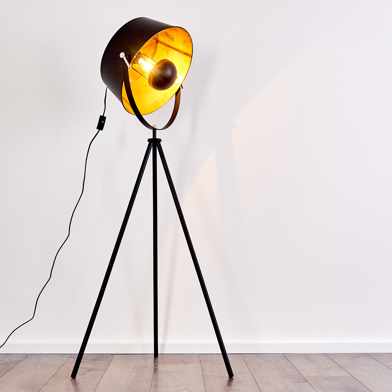 Eine schwarze Stehlampe mit goldenem Innenschirm steht auf einem Holzboden vor einer weißen Wand.