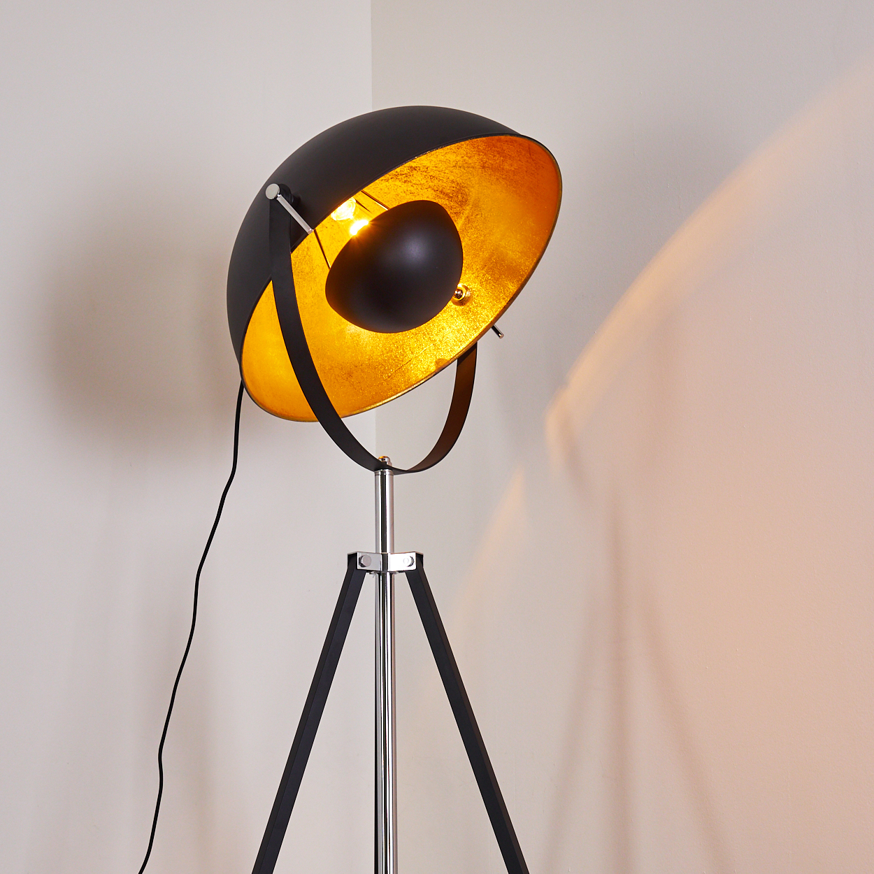 Eine schwarz-goldene Stehlampe mit Dreibein. Die Lampe ist an und wirft ein warmes Licht an die Wand. Ein schwarzes Kabel ist sichtbar.