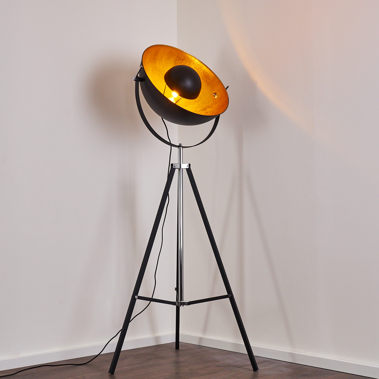 Eine schwarz-goldene Stehlampe mit Dreibein-Sockel. Die Lampe ist eingeschaltet und wirft ein warmes Licht. Hintergrund ist eine weiße Wand und Holzfußboden.