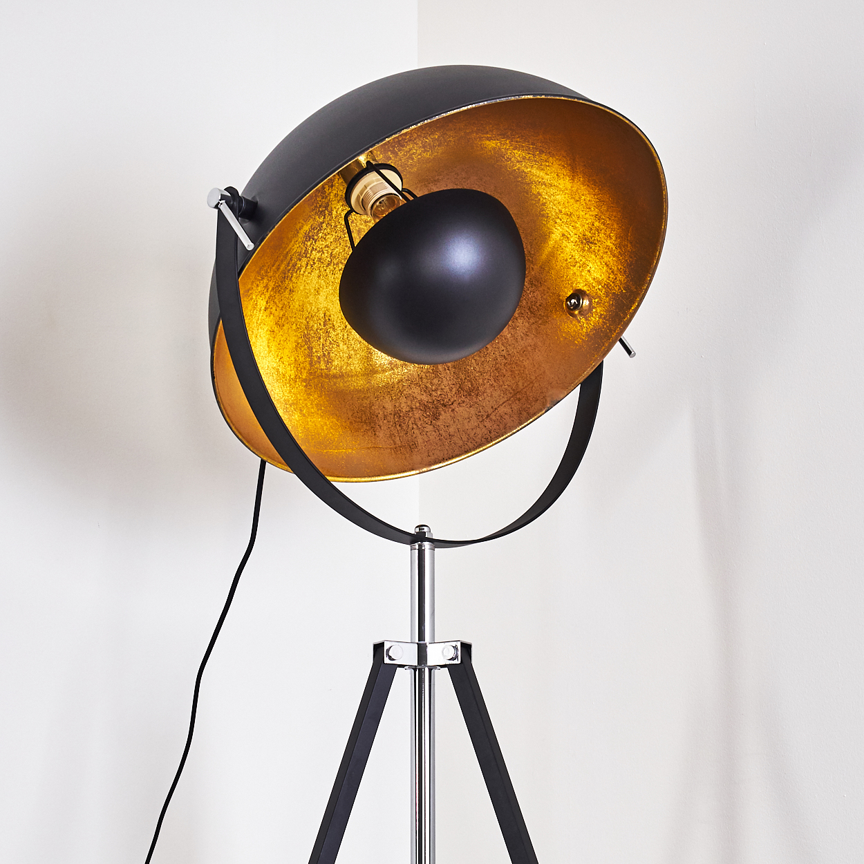 Eine moderne schwarze und goldene Stehlampe mit Dreibein-Sockel. Die Lampe hat einen großen, runden Schirm und ein schwarzes Kabel.