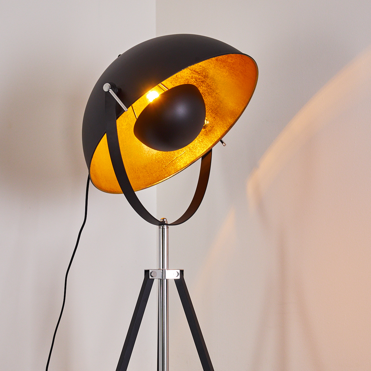 Moderne schwarz-goldene Stehlampe mit Dreibein-Sockel. Das Innere der Lampe ist goldfarben. Die Lampe ist an und wirft Licht.