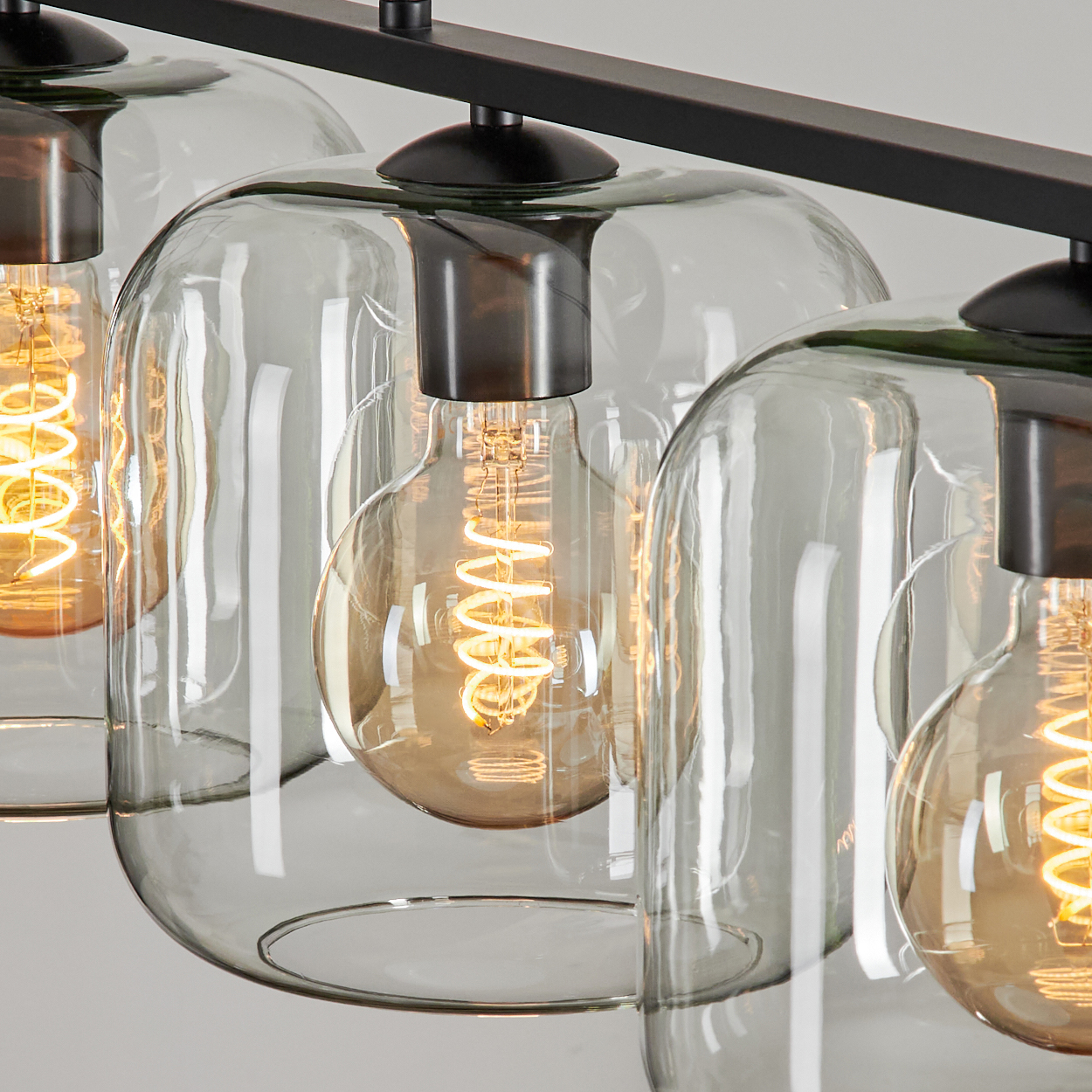 Nahaufnahme einer modernen Leuchte. Sie hat klare Glasschirme, Edison-Glühbirnen und einen dunklen Metallrahmen vor weißem Hintergrund.