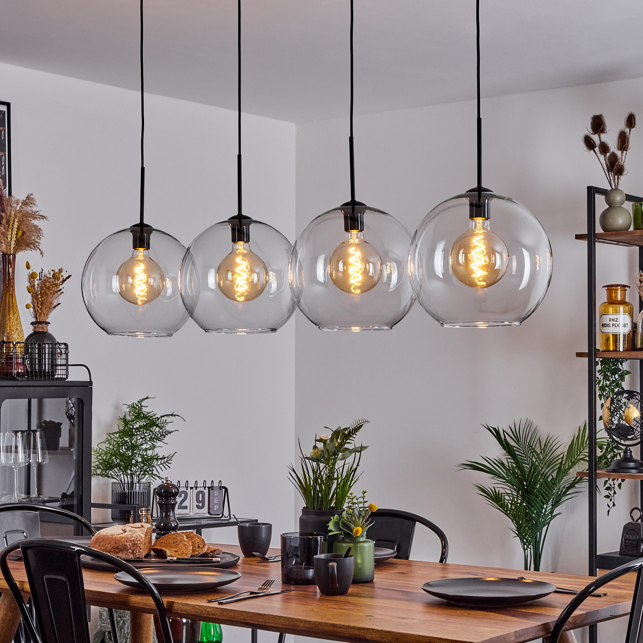 Esszimmer mit Holztisch, schwarzen Stühlen, vier kugelförmigen Pendelleuchten. Tisch gedeckt mit Tellern, Brot und Pflanzen.