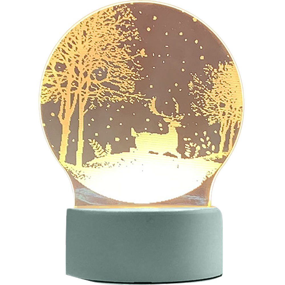 Eine runde Lampe mit einer Waldszene und einem Hirsch. Der Sockel ist hellblau. Der Wald wird von einem warmen Schein mit Schnee beleuchtet.