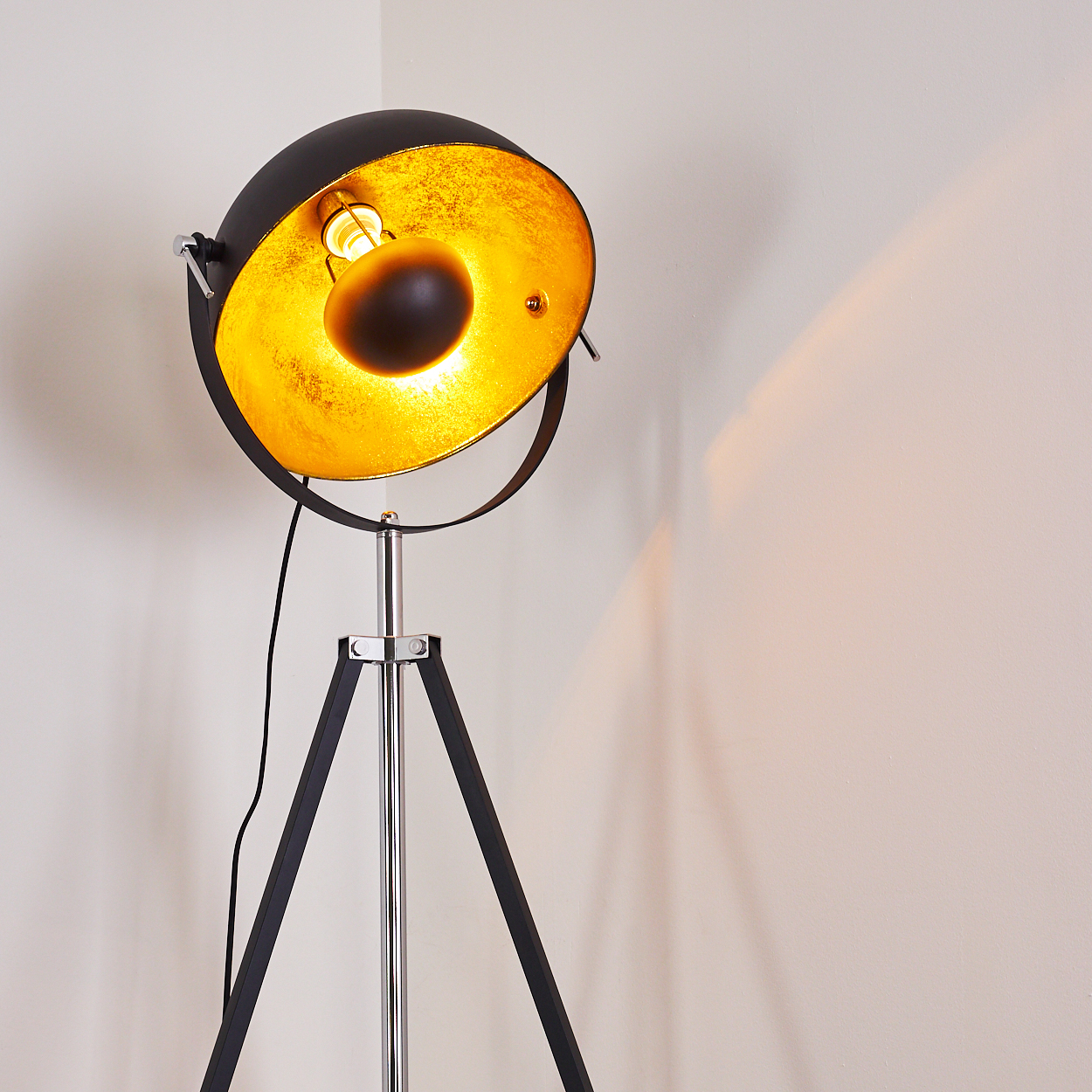 Eine schwarz-goldene Stehlampe mit Dreibein-Sockel. Die Lampe ist eingeschaltet und wirft Licht auf die Wand.